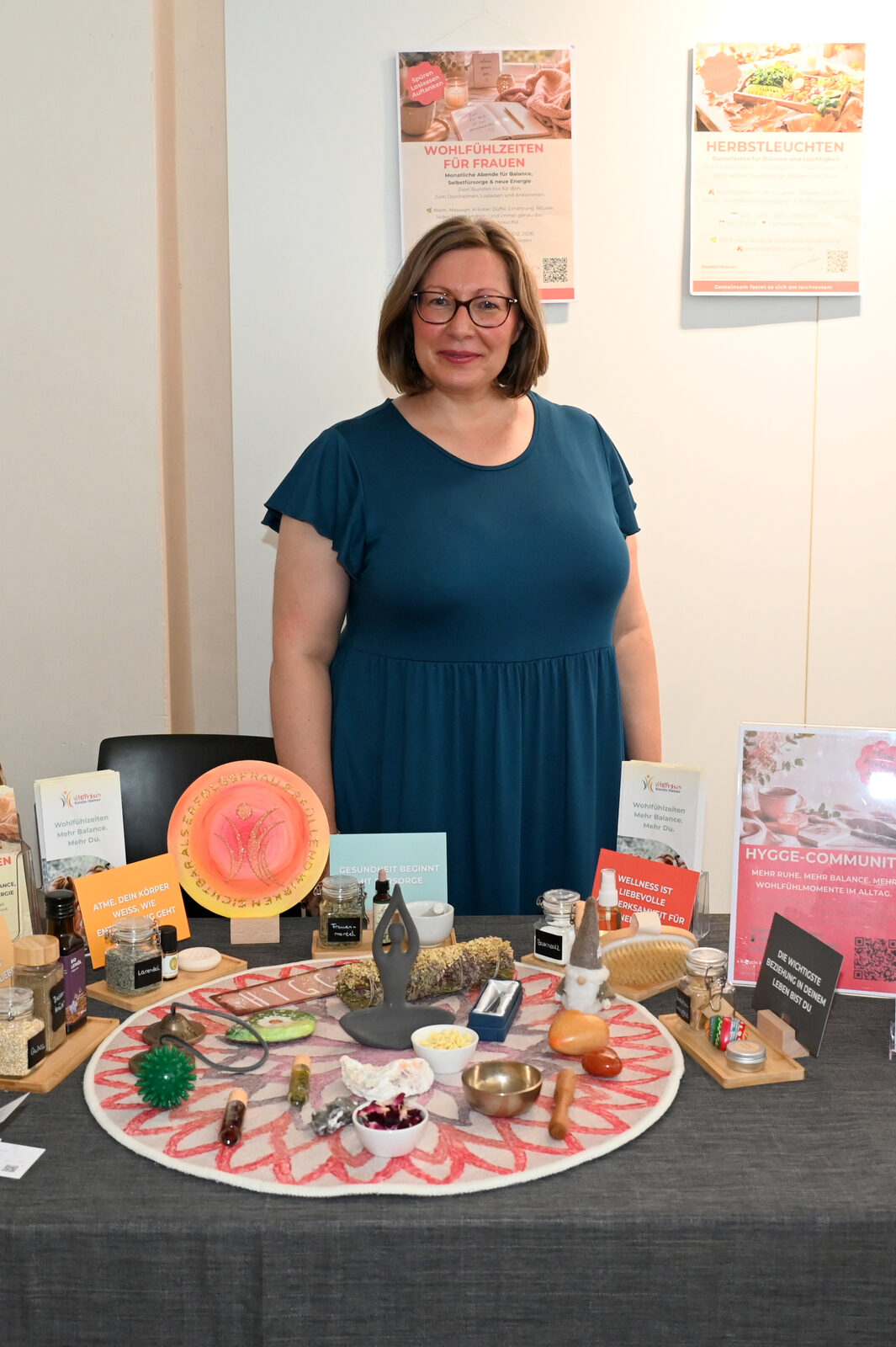 Kerstin Hiemer steht lächelnd an ihrem Messestand mit Wohlfühlzeiten- und Herbstleuchten-Postern im Hintergrund